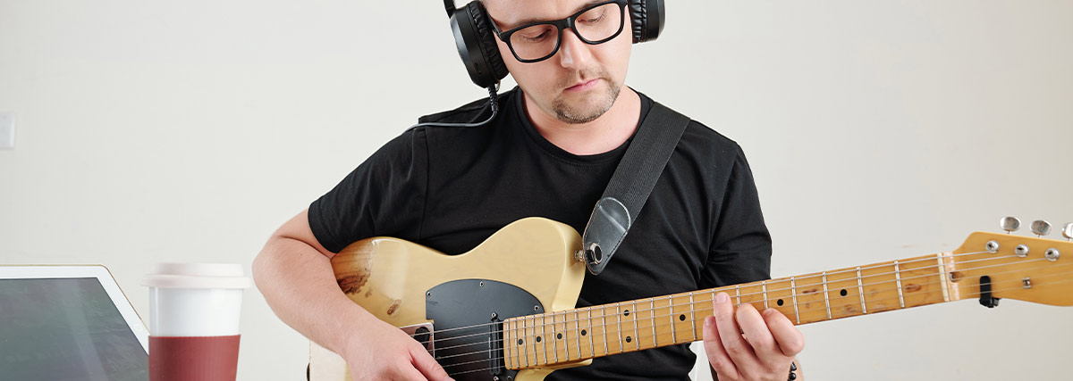 Adult guitarist practising electric guitar with headphones, sheet music and laptop